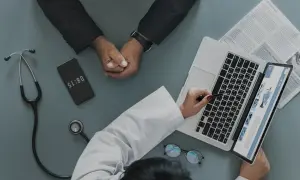 Médico revisa informe en laptop, paciente espera. El teléfono marca 08:15, quizás para justificar faltar al trabajo sin justificante médico.