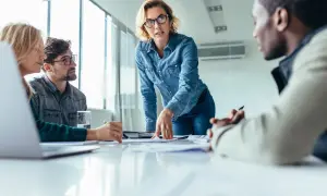 Mujer con gafas dirige reunión, explicando un proyecto. La comunicación vertical fluye en este equipo.