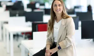 Mujer sonriente en oficina moderna, sentada en un escritorio. Su atuendo profesional y su actitud reflejan el valor añadido de una empresa.