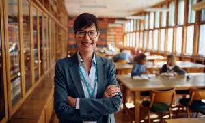 Profesora sonriente en biblioteca, lista para el test de gestión procesal. Estudiantes estudian al fondo.