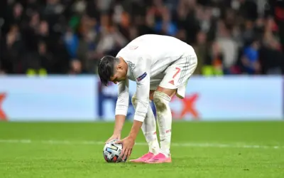 Jugador de fútbol español se agacha para patear un balón, reflexionando sobre cómo se calcula el resultado en penaltis dinero.