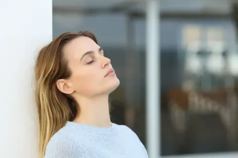 Mujer con ojos cerrados, apoyada en pared blanca, reflexionando sobre cuanto se cobra estando de baja por ansiedad.