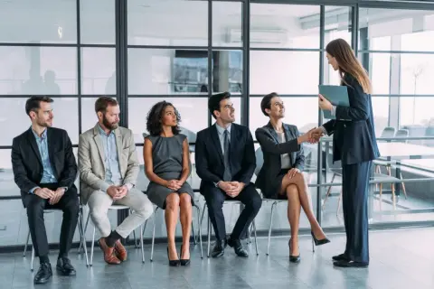 Un grupo de personas espera en fila para una entrevista de trabajo. Una mujer sonríe mientras le da la mano a otra mujer, quizás sintiendo que no me adapto a mi nuevo trabajo.
