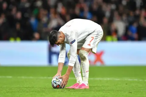 Jugador de fútbol español se agacha para patear un balón, reflexionando sobre cómo se calcula el resultado en penaltis dinero.