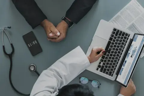 Médico revisa informe en laptop, paciente espera. El teléfono marca 08:15, quizás para justificar faltar al trabajo sin justificante médico.