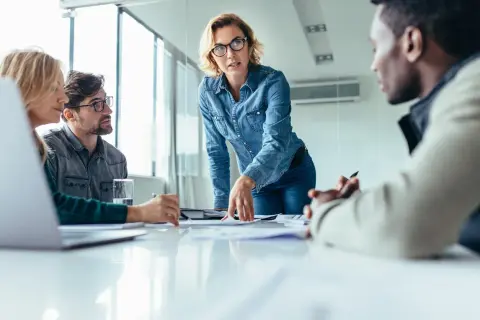 Mujer con gafas dirige reunión, explicando un proyecto. La comunicación vertical fluye en este equipo.
