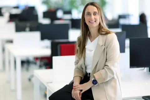 Mujer sonriente en oficina moderna, sentada en un escritorio. Su atuendo profesional y su actitud reflejan el valor añadido de una empresa.