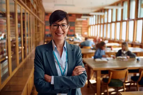 Profesora sonriente en biblioteca, lista para el test de gestión procesal. Estudiantes estudian al fondo.