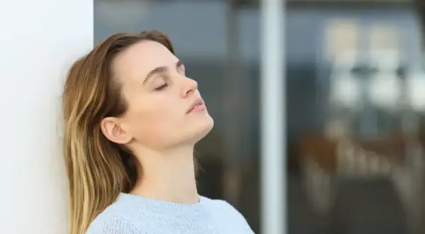 Mujer con ojos cerrados, apoyada en pared blanca, reflexionando sobre cuanto se cobra estando de baja por ansiedad.
