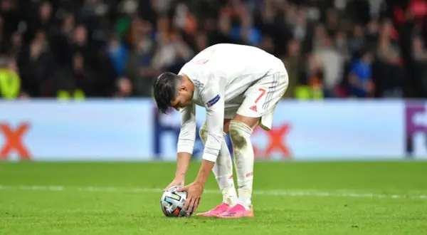 Jugador de fútbol español se agacha para patear un balón, reflexionando sobre cómo se calcula el resultado en penaltis dinero.