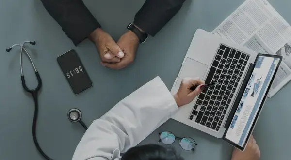 Médico revisa informe en laptop, paciente espera. El teléfono marca 08:15, quizás para justificar faltar al trabajo sin justificante médico.