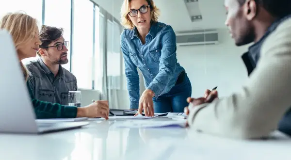 Mujer con gafas dirige reunión, explicando un proyecto. La comunicación vertical fluye en este equipo.