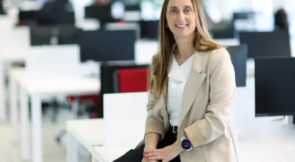 Mujer sonriente en oficina moderna, sentada en un escritorio. Su atuendo profesional y su actitud reflejan el valor añadido de una empresa.