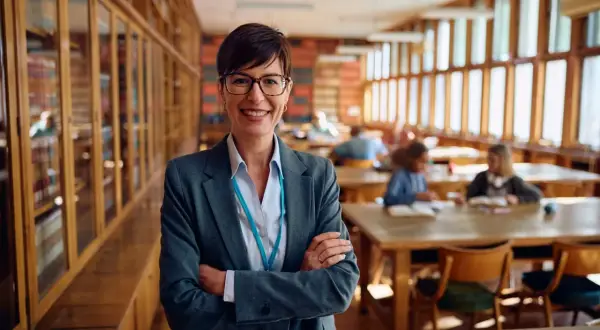 Profesora sonriente en biblioteca, lista para el test de gestión procesal. Estudiantes estudian al fondo.