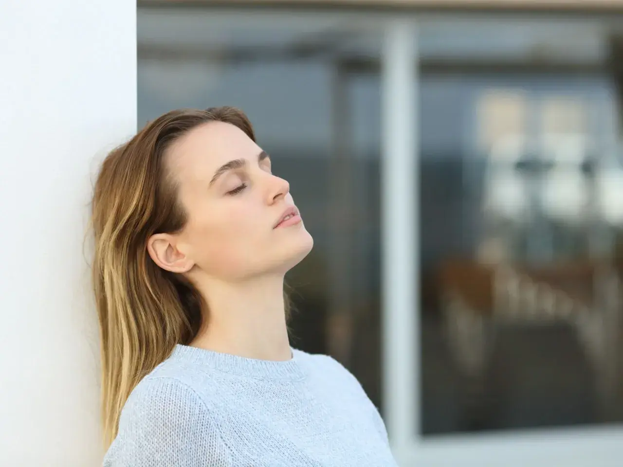 Mujer con ojos cerrados, apoyada en pared blanca, reflexionando sobre cuanto se cobra estando de baja por ansiedad.