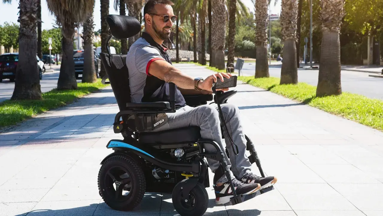 Hombre sonriente en silla de ruedas eléctrica, con un teléfono en la mano, paseando por un camino arbolado. Se puede trabajar con una discapacidad del 65%.