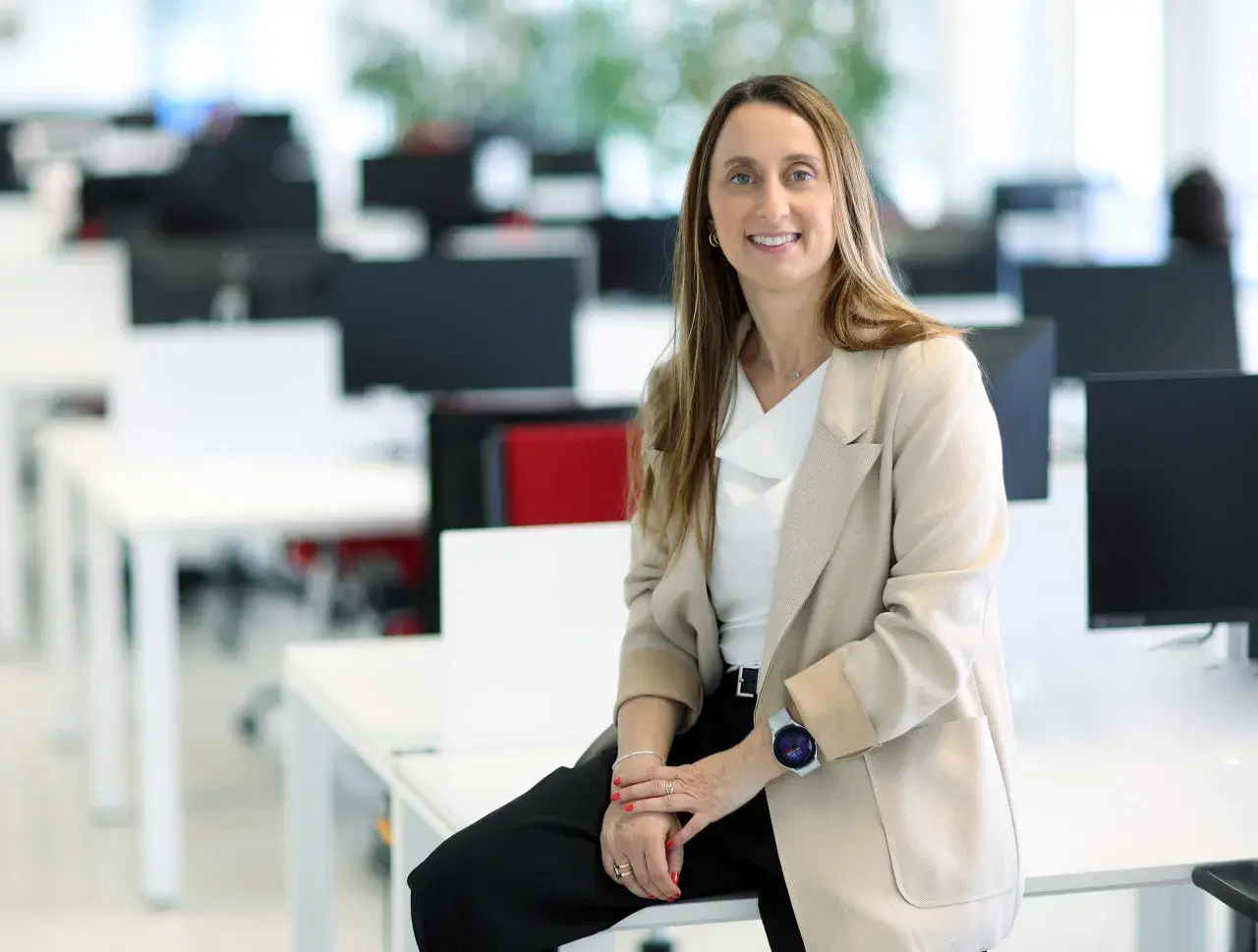 Mujer sonriente en oficina moderna, sentada en un escritorio. Su atuendo profesional y su actitud reflejan el valor añadido de una empresa.