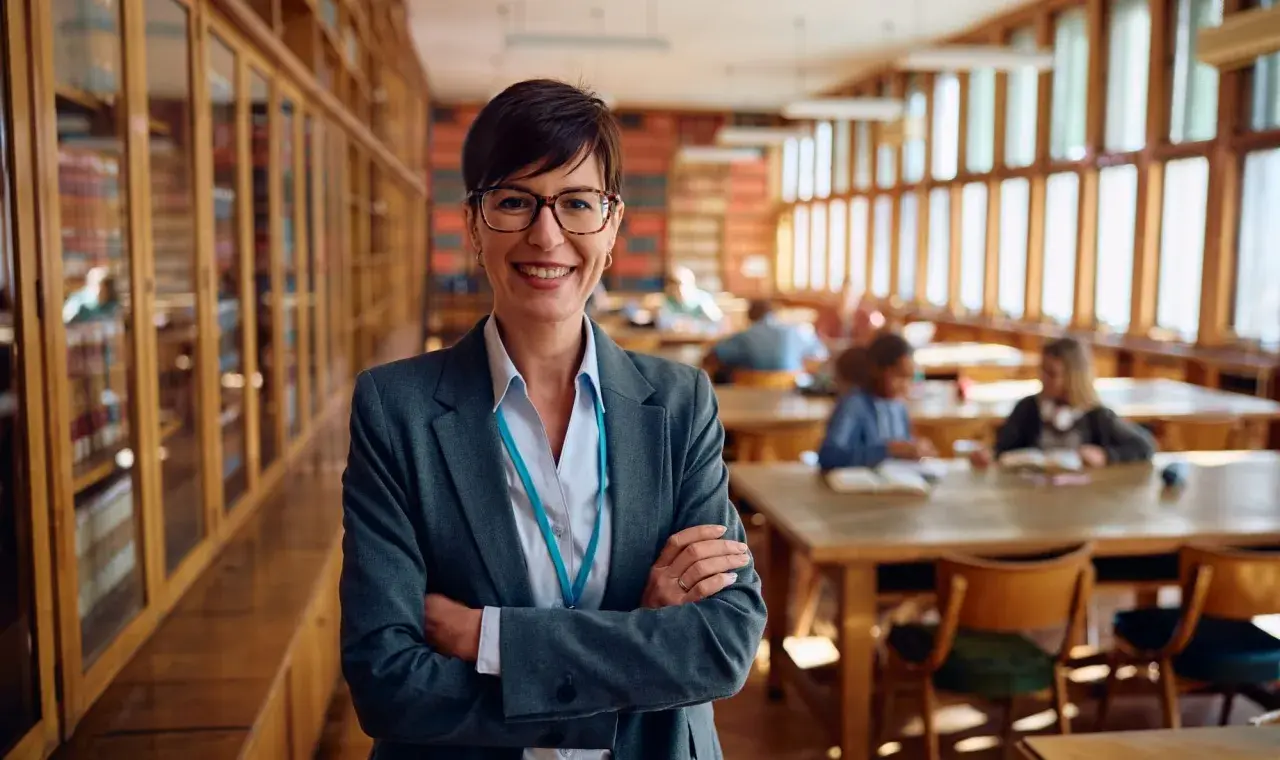 Profesora sonriente en biblioteca, lista para el test de gestión procesal. Estudiantes estudian al fondo.