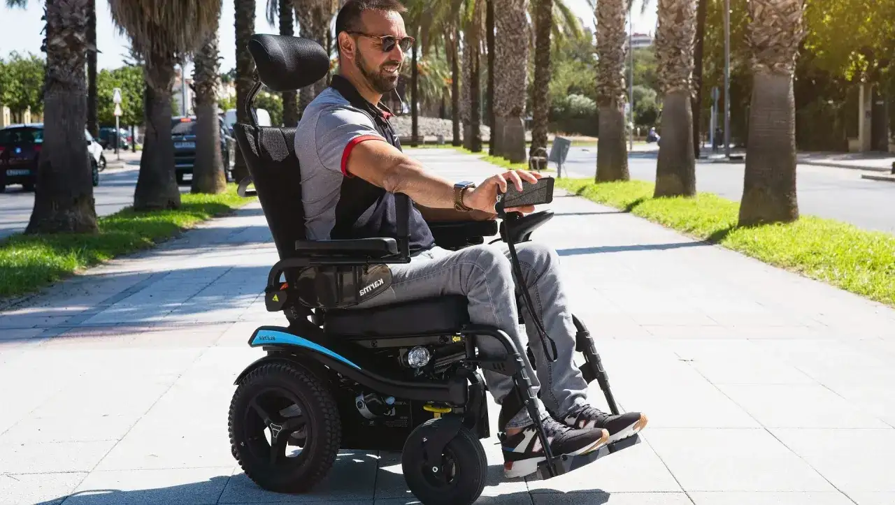 Hombre sonriente en silla de ruedas eléctrica, con un teléfono en la mano, paseando por un camino arbolado. Se puede trabajar con una discapacidad del 65%.