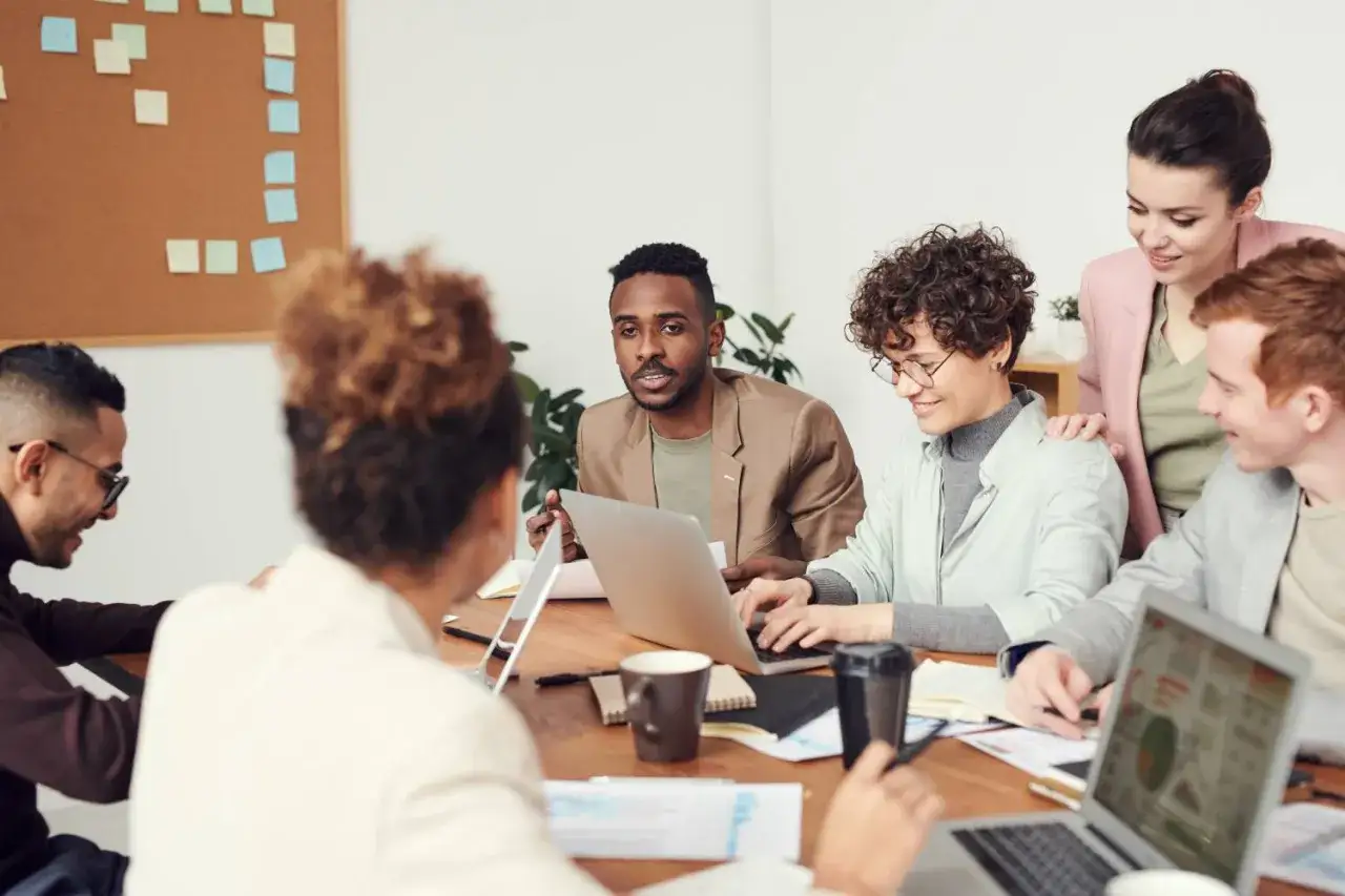 Un equipo diverso colabora en una reuni&oacute;n, discutiendo ideas. Un hombre con chaqueta marr&oacute;n es un business partner clave, mientras otros analizan gr&aacute;ficos en laptops.