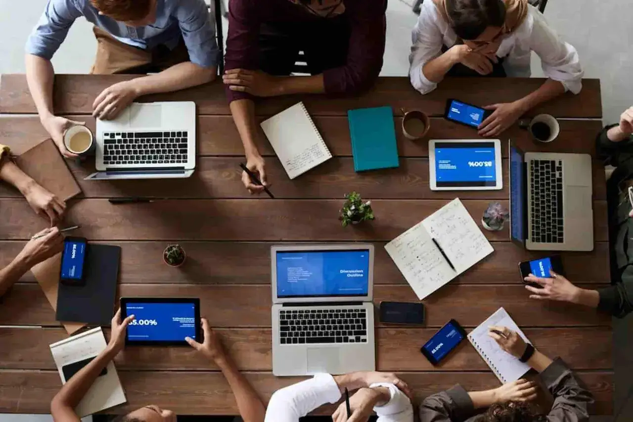 Equipo colaborando en una mesa de madera, con laptops, tablets y tel&eacute;fonos. La comunicaci&oacute;n laboral fluye mientras toman notas y revisan informaci&oacute;n.