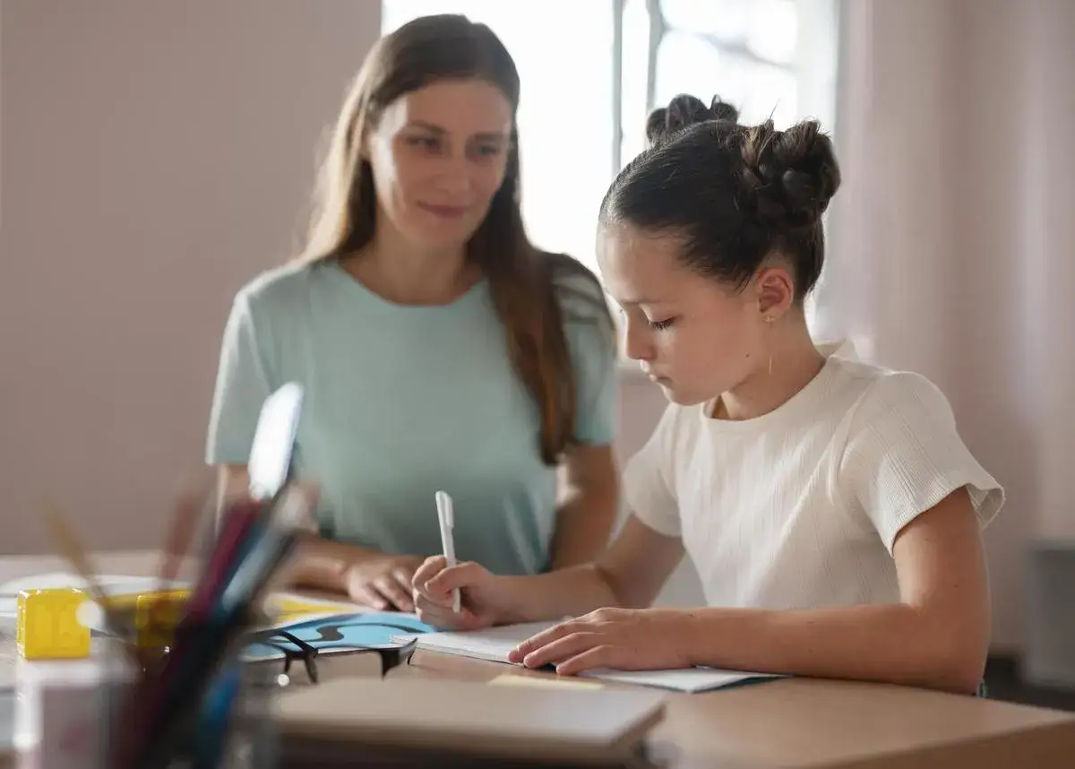 Niña concentrada escribiendo, con una mujer observándola. Una escena de apoyo en la oposición orientación educativa.