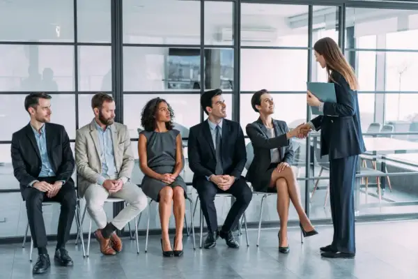 Un grupo de personas espera en fila para una entrevista de trabajo. Una mujer sonríe mientras le da la mano a otra mujer, quizás sintiendo que no me adapto a mi nuevo trabajo.