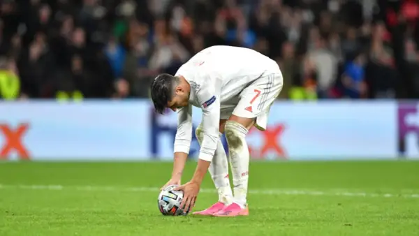 Jugador de fútbol español se agacha para patear un balón, reflexionando sobre cómo se calcula el resultado en penaltis dinero.
