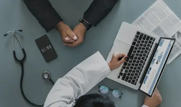 Médico revisa informe en laptop, paciente espera. El teléfono marca 08:15, quizás para justificar faltar al trabajo sin justificante médico.